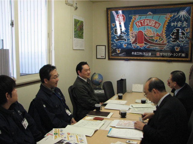 ネイチャーテック釧路の事務所で社長（左列奥）らの話を聞くプラスチック処理促進協会調査部（右列）。正面の大漁旗は同社創立のきっかけを作ったガラスリソーシング（株）から贈呈された。
